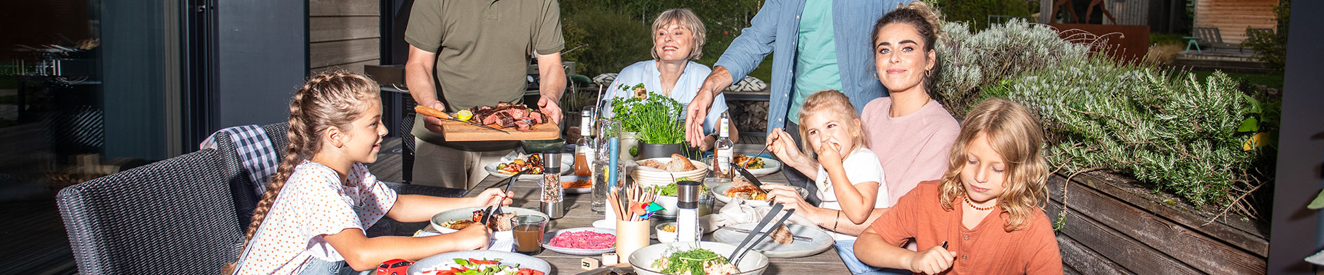 Familie sitzt zusammen mit Kindern am Tisch und Mann serviert gegrilltes Essen.