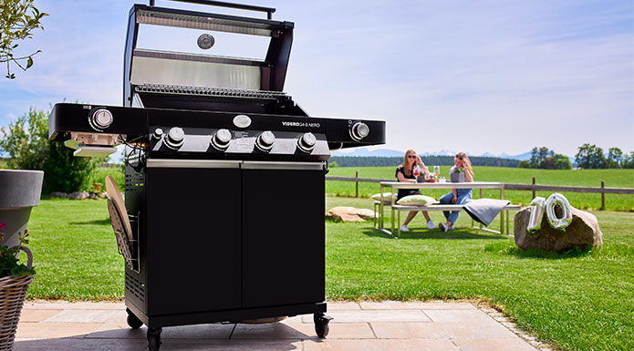 Gasgrill mit geöffnetem Deckel im Vordergrund. Dahinter zwei Damen, die mit Weingläser anstoßen.