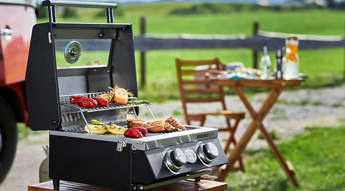 RÖSLE Gasgrill BBQ-Portable Videro G2-P Videro G2-P mit geöffnetem Deckel und diversem Grillgut im Vordergrund, dahinter kleiner Tisch und Stuhl.